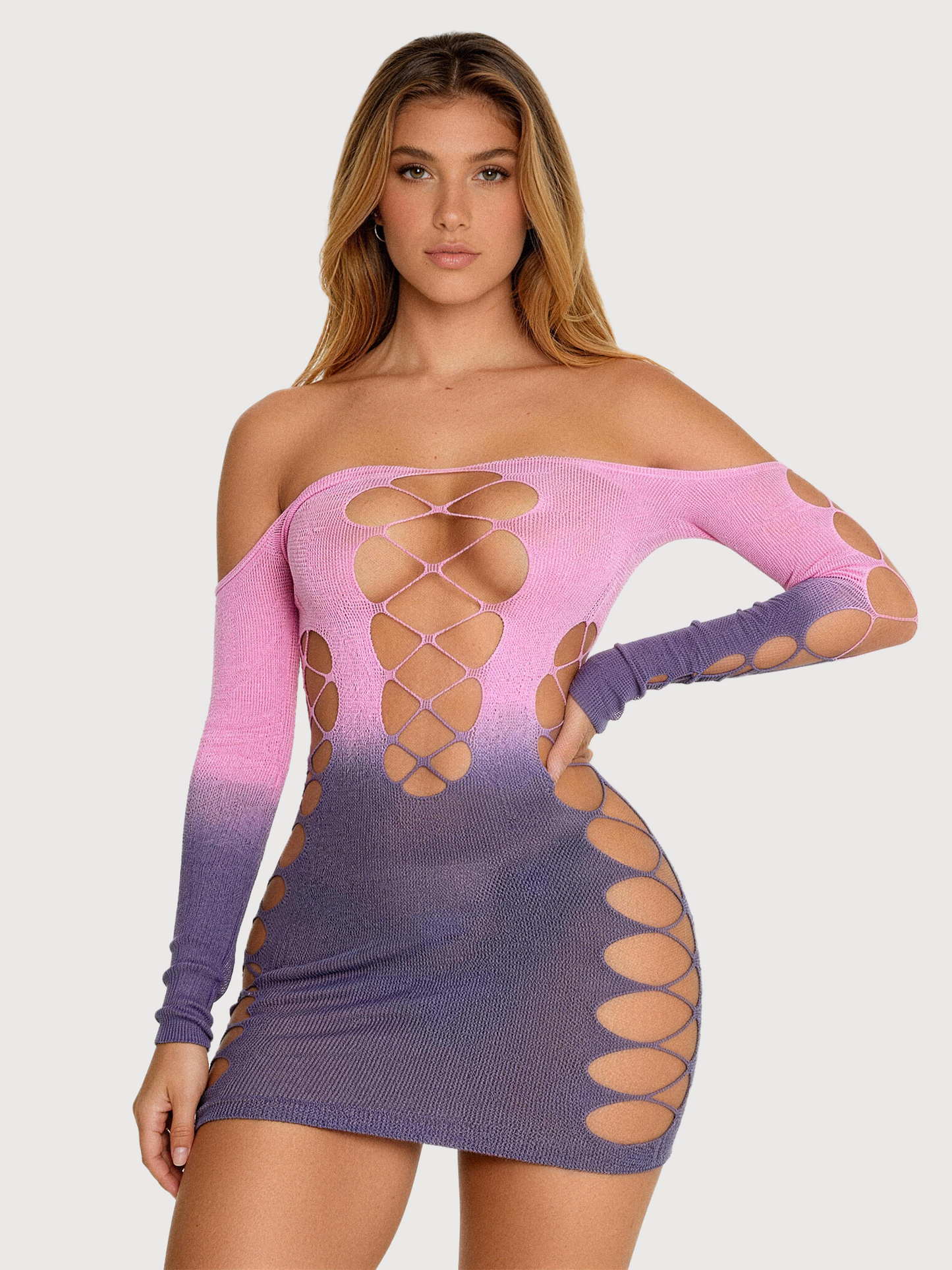 A woman modeling a long-sleeve off-the-shoulder knit mini dress with a pink-to-purple ombré gradient and large cutout details on the bodice, arms, and hips, against a light gray studio background.