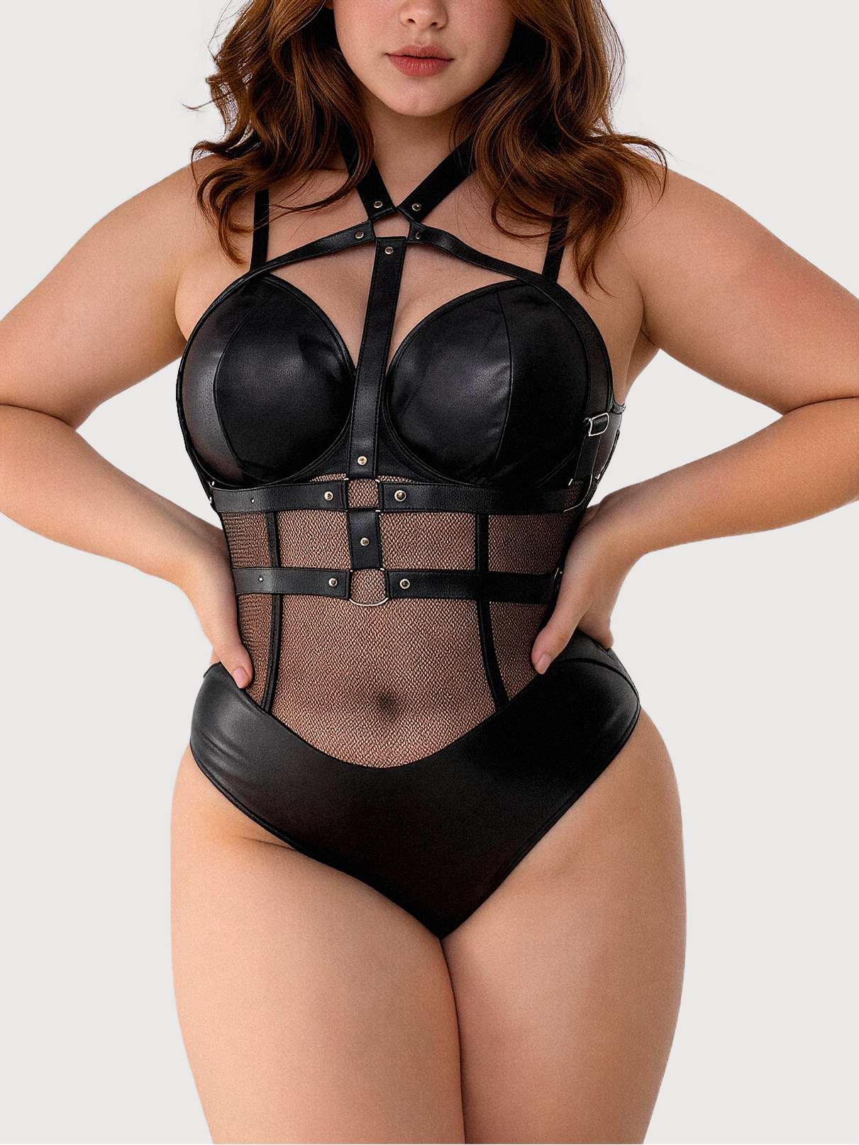A woman modeling a black strappy lingerie bodysuit with faux leather-like panels and sheer mesh, photographed against a plain light gray studio background.