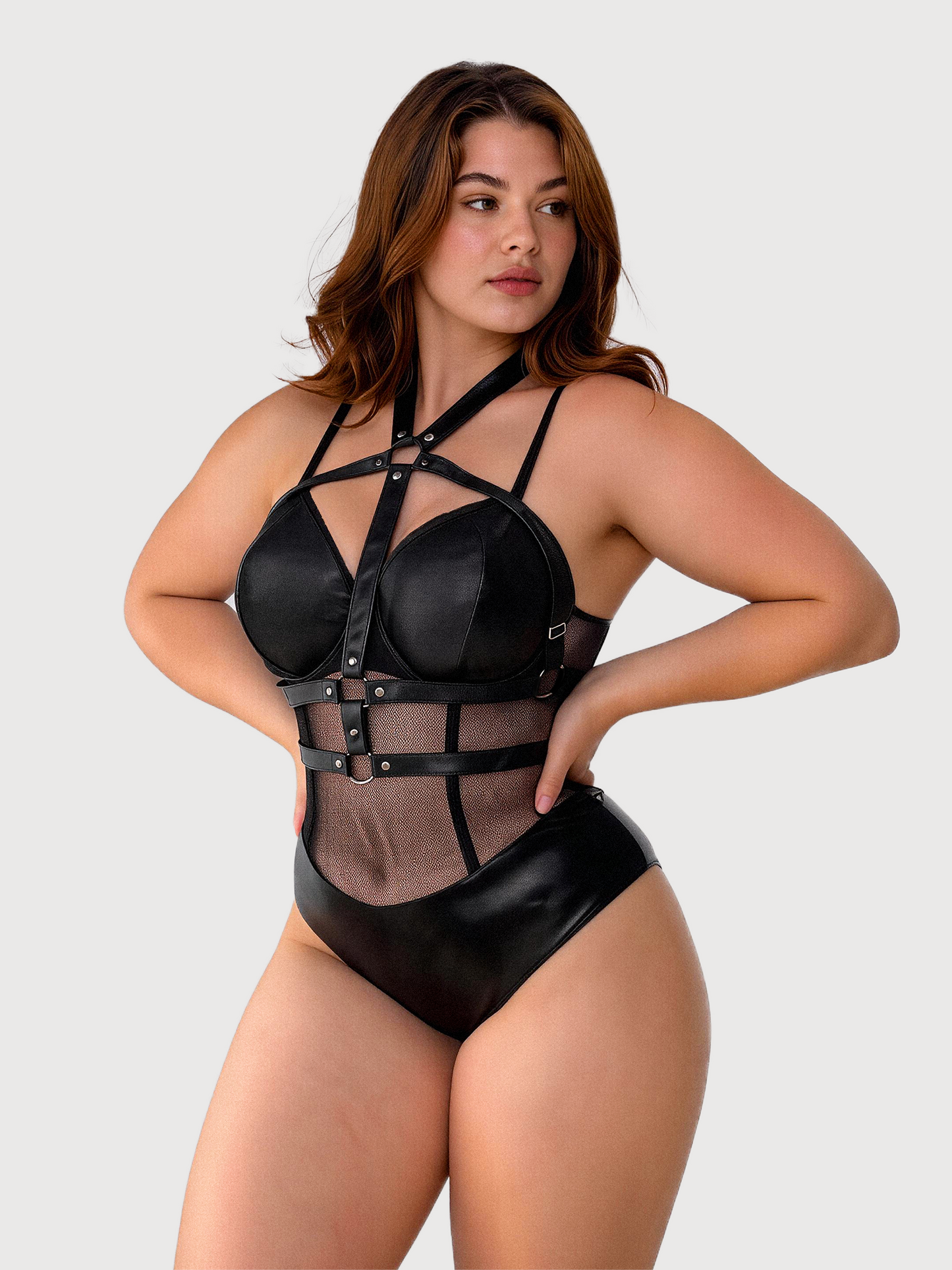 A woman modeling a black strappy lingerie bodysuit with sheer mesh panels and silver-tone hardware, photographed against a plain light gray studio background.