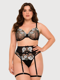 A plus-size model wearing a black lace lingerie set with white floral embroidery, including a bra, high-waisted briefs, and garter straps, against a light gray background.