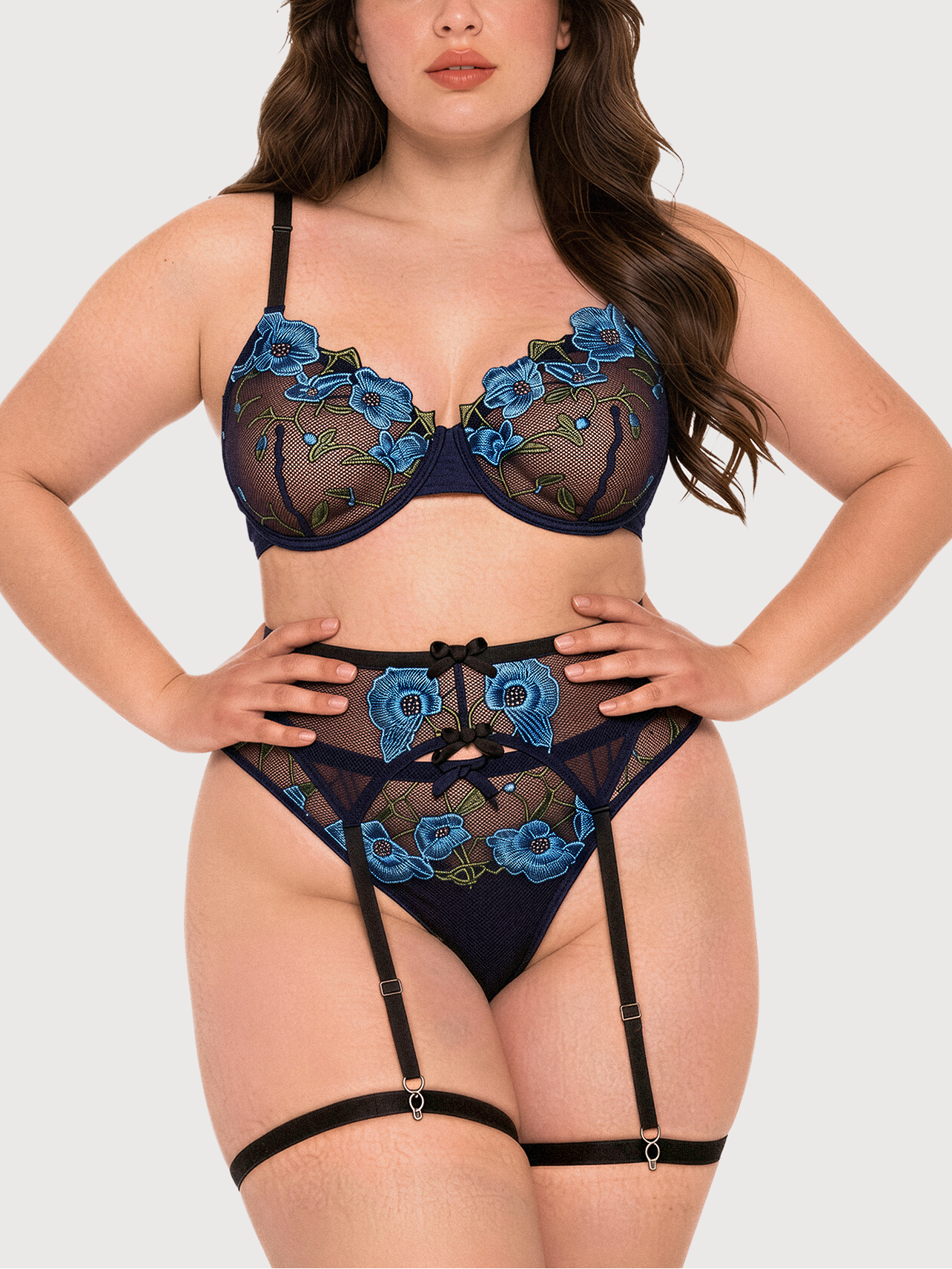 A woman modeling a three-piece lingerie set featuring a sheer black mesh bra and panty with blue floral embroidery, paired with a matching garter belt and black thigh bands, against a solid light gray background.