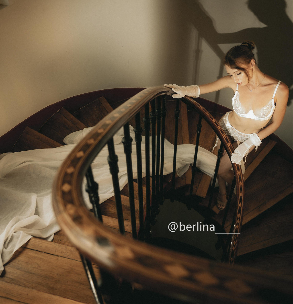A woman in a white lingerie set and white gloves sits on the steps of a wooden spiral staircase, looking down. The scene is shot from an elevated angle with warm, moody lighting and a draped white fabric on the floor.