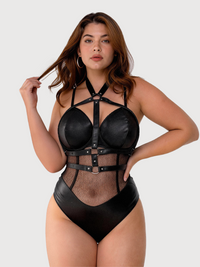 A woman modeling a black strappy one-piece bodysuit with faux leather-like panels and sheer mesh sections, photographed against a light gray studio background.