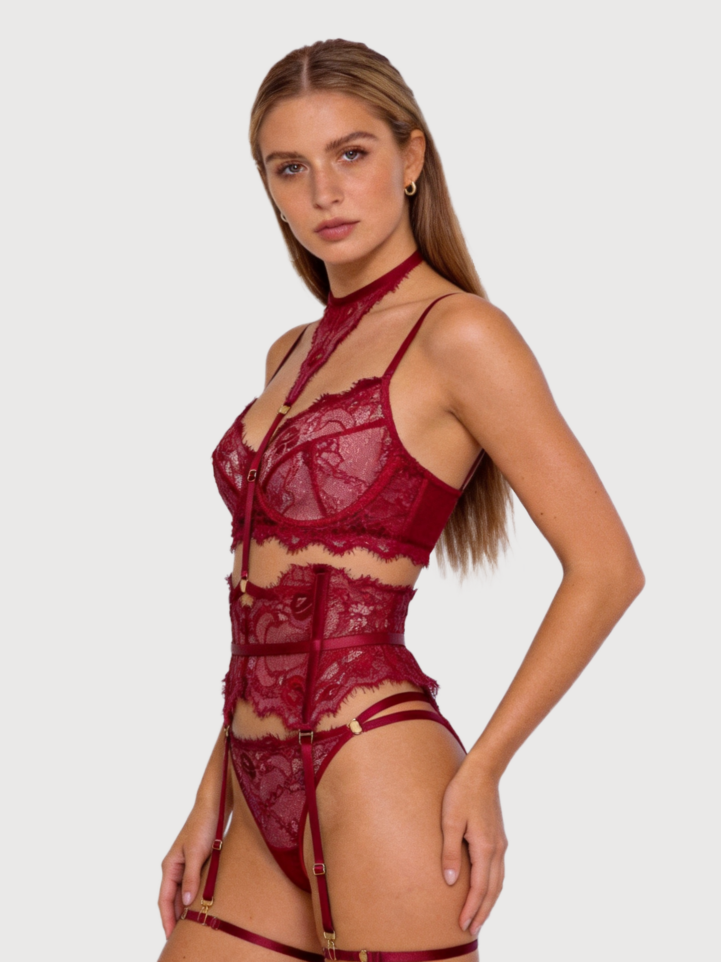 A woman modeling a deep burgundy lace lingerie set with a high-neck choker, sheer cups, side cutouts, a structured corset-style bodice, matching garter belt, and panty, photographed against a solid light gray background.