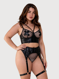 A woman modeling a black strappy lingerie set with faux leather and fishnet mesh details against a light gray studio background.