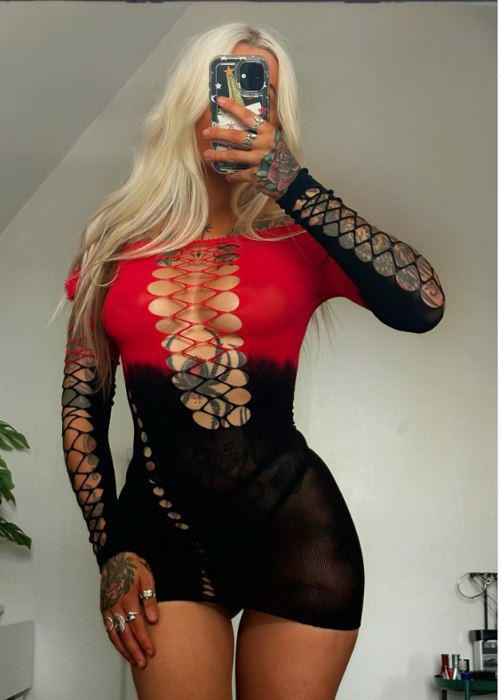 A woman takes a mirror selfie wearing a sheer long-sleeve bodysuit with a red-to-black gradient and cutout lace-up details on the front. The photo is taken in a home setting with a plant and wall visible in the background.