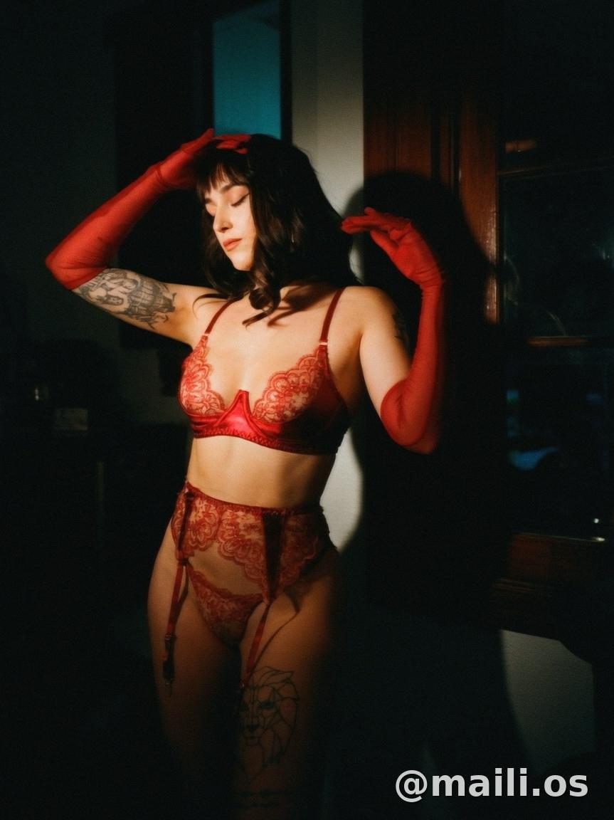 A woman modeling a red lace lingerie set with matching red gloves in a dimly lit room. The set includes a lace bra and coordinating panty with garter details.